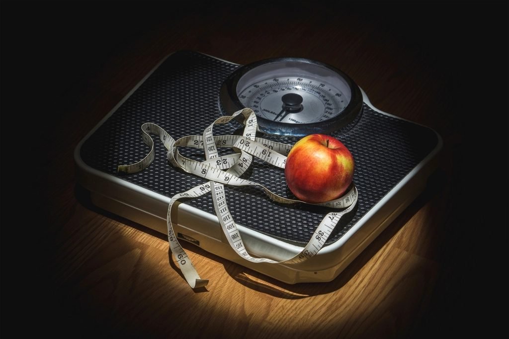 Measuring tape wrapped around a green apple beside a bathroom scale, representing weight and calorie tracking