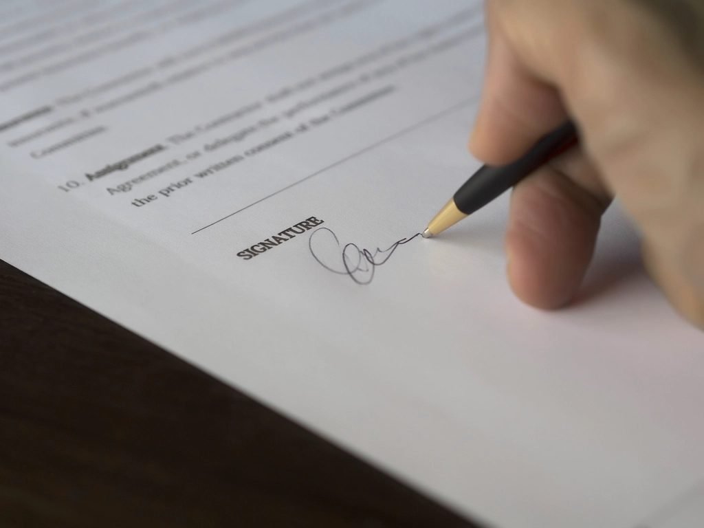 Person signing a document by hand next to a laptop, representing electronic signature workflows